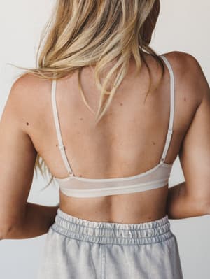 A back-facing shot highlighting the delicate spaghetti straps and sheer mesh panel of a nude bralette, paired with a smocked light denim skirt — soft, understated, and effortlessly wearable.