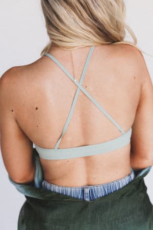 A woman with long blonde hair is shown from behind, wearing a sage Crochet High Neck Bralette and high-waisted jeans, with a green jacket draped around her arms—an effortless layering piece.