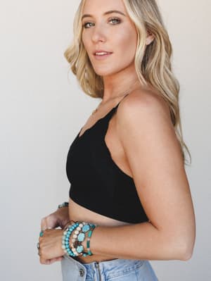 A blonde model shot in three-quarter profile against a soft neutral studio wall, wearing a black cropped bralette with a stacked collection of turquoise and stone beaded bracelets as the clear focal point — casual, expressive, effortlessly boho.