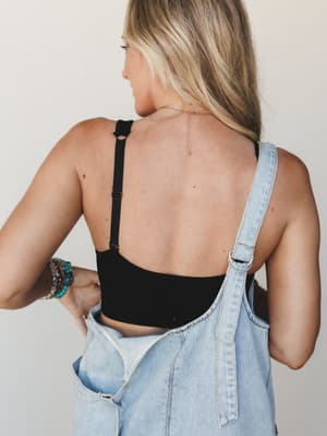 Shot from behind, a model pairs a black bralette with light-wash denim overalls, the open back and delicate gold chain necklace doing quiet, effortless work — stacked beaded bracelets on her wrist adding just the right amount of personality.