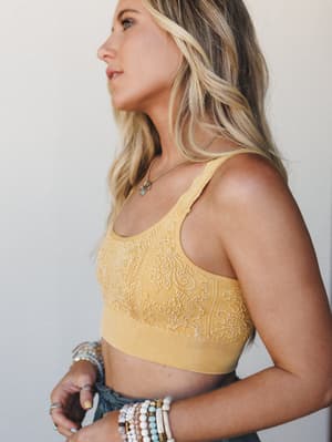 A sun-warmed side profile shot of a model in a textured mustard bralette with tonal embroidery, layered with beaded bracelets and a delicate pendant necklace — the kind of effortless warm-weather outfit that looks like it was put together in about tw