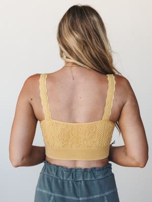 A back-facing shot of a textured mustard bralette with intricate floral jacquard detailing and lace-edged straps, paired with washed slate wide-leg pants — soft studio light keeps the focus on the fabric texture and open back.