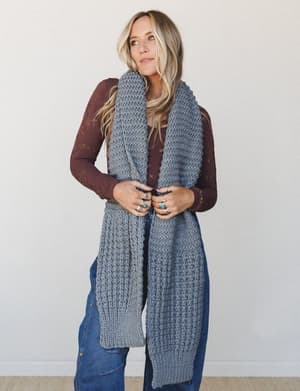 A relaxed, earthy layered look — a chunky knit oversized gray scarf worn long over a burgundy pointelle long-sleeve top and dark wash flared jeans, styled with stacked silver and turquoise rings for that collected, intentional boho feel.