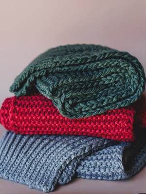 A cozy stack of three chunky knit sweaters folded together — forest green on top, cranberry red in the middle, and dusty blue-gray at the base — shot close against a soft blush background with a still-life, tactile warmth to it.