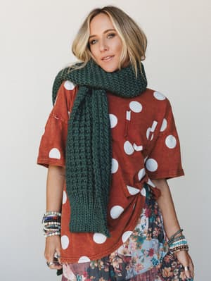A layered, lived-in look built around a chunky knit scarf in deep forest green, worn over a rust polka dot oversized top and a peeking floral midi skirt — stacked bracelets on both wrists and the whole thing feels like a really good Saturday.