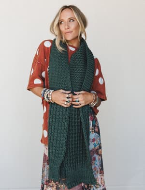 A layered fall look built around an oversized chunky knit scarf in deep forest green, worn over a rust polka dot top and a floral maxi skirt — stacked bracelets and statement rings pull the whole collected-over-time feel together.