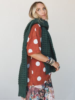 A layered, expressive look — chunky forest green knit scarf draped over a rust polka dot tee and a floral tiered skirt, styled with a stack of beaded bracelets and a gaze that says she's already somewhere else.