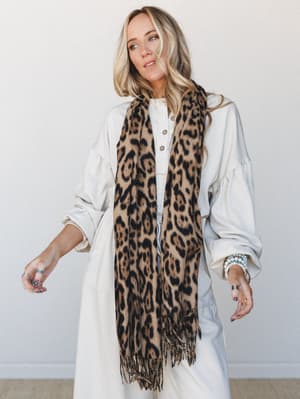 A long leopard print scarf with fringe ends draped over a cream oversized dress — the kind of effortless layering that makes a neutral outfit feel like it had a plan all along.