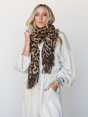 A relaxed, effortlessly styled shot of a model in a cream zip-up jumpsuit layered with an oversized camel leopard print scarf, accessorized with turquoise and pearl bracelets and a statement ring — the kind of outfit that looks like you tried just th