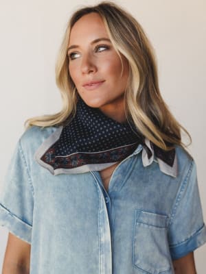 A soft, effortless mid-shot of a model in a light wash chambray button-down wearing a navy bandana scarf tied at the neck — the kind of finishing touch that makes a simple outfit feel completely intentional.