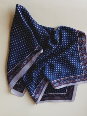 A navy bandana scarf folded loosely on a cream surface, showing off its diamond ditsy print center and a rich burgundy paisley border — the kind of accessory that quietly makes an outfit.