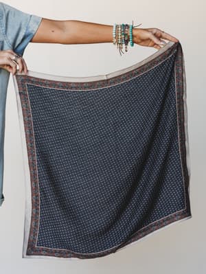 A model holds out a navy square scarf with a small ditsy print and a rust and sage floral border, styled with layered turquoise and beaded bracelets and a chambray shirt — the kind of accessory that does five things and takes up no space in your bag.