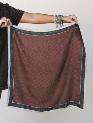 A model holds up a large square bandana scarf in rust with a ditsy floral print and navy paisley border, styled against a neutral wall with a stacked bracelet arm adding a lived-in boho touch.