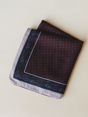 A folded bandana scarf laid flat, showing a rich burgundy center with tiny geometric diamond print, bordered by a deep navy floral edge and finished with a soft lavender-grey hem — the kind of piece that looks equally at home tucked into a bag or tie