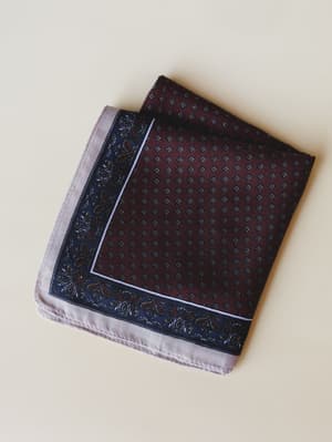 A folded bandana scarf laid flat, showing a rich burgundy center with tiny geometric diamond print, bordered by a deep navy floral edge and finished with a soft lavender-grey hem — the kind of piece that looks equally at home tucked into a bag or tie