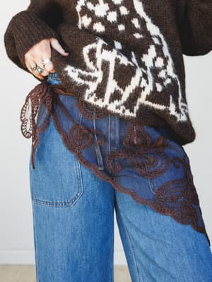 A mid-shot styling detail of a sheer brown lace handkerchief tied at the hip over wide-leg medium-wash jeans, layered beneath a chunky dark brown animal-print knit sweater — the kind of layering trick that makes an outfit feel collected and a little