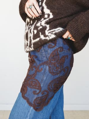 A close-up detail shot of wide-leg denim jeans with bold brown paisley embroidery at the hip, paired with a chunky dark brown fair isle sweater and stacked silver and turquoise rings — the kind of outfit that feels like it was pulled from a really go
