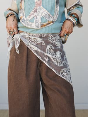 A mid-shot styled look centered on a sheer lace paisley handkerchief tied at the hip over wide-leg brown trousers, layered with a patchwork vest and soft blue thermal — the kind of outfit that looks like it came together effortlessly but took real in