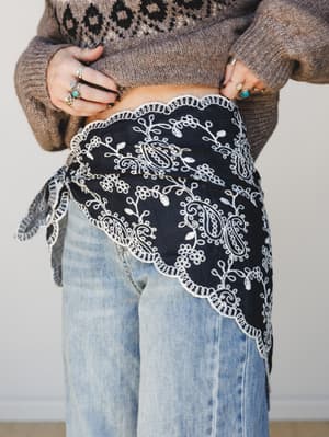 A black embroidered handkerchief wrap tied at the hip over light wash wide-leg jeans and a fair isle sweater — the kind of layered, personal styling that makes a simple outfit feel completely intentional.