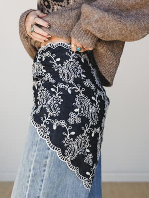 A charcoal bandana-style handkerchief with white paisley embroidery and scalloped trim tucked into light wash jeans, styled with a heathered brown knit sweater and layered silver and turquoise rings — the kind of small detail that makes the whole out
