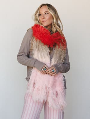 A model layers a shaggy ombre faux fur scarf — graduating from red-orange at the collar through cream and into blush pink — over a soft taupe long-sleeve top, styled with pink striped wide-leg pants and stacked turquoise and silver rings for a playfu