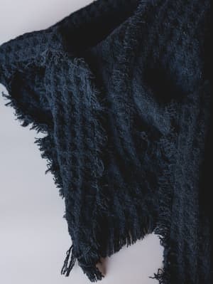 A close-up detail shot of a deep navy-black textured scarf with a chunky boucle-like weave and delicate fringe trim along the edges, draped softly against a light gray studio surface.
