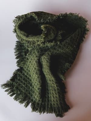 A deeply textured olive green scarf loosely knotted and styled on a clean white surface — the kind of cozy accessory that feels like it belongs draped over a chunky sweater on a slow autumn morning.