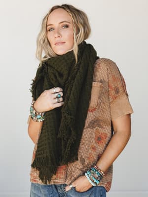 A relaxed, layered look built around an oversized olive fringe scarf draped over a faded boho-print tee — the kind of outfit that looks effortless but clearly wasn't an accident.