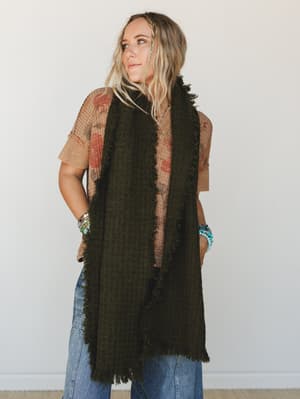 A long olive boucle scarf styled as a vest over a floral waffle-knit tee and wide-leg denim, accessorized with turquoise and beaded bracelets — the kind of layered, lived-in look that feels effortless without trying.