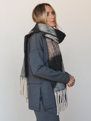 A blonde model shot in profile wears an oversized color-block scarf in cream, grey, and black draped over a slate blue longline top and matching pants — the kind of cozy, pulled-together look that makes a Tuesday feel intentional.