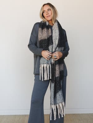 A soft, relaxed outfit anchored by an oversized plaid scarf with cream fringe — styled over a slate blue matching set for that effortless 'grabbed the coziest thing on the way out' energy.