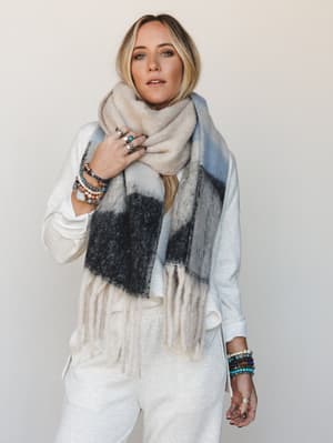 A relaxed, effortlessly styled shot of a model wrapped in an oversized color-block scarf with fringe ends, layered over a cream sweatshirt and joggers — the kind of cozy-cool outfit you'd wear on a slow Saturday and still feel put-together.