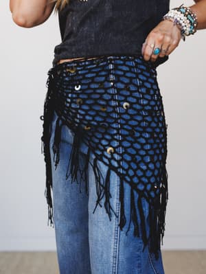 A close-up of a black macramé fringe hip belt layered over medium-wash denim jeans and a charcoal tee, styled with stacked stone and turquoise bracelets and boho rings — the kind of piece that turns a basic outfit into a whole mood.