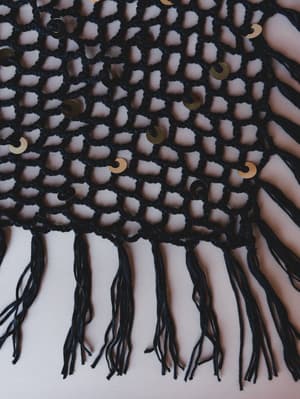 A close-up detail shot of a black open-weave crochet piece scattered with gold sequin embellishments and finished with long, loose fringe trim — the kind of piece that looks like it belongs at a bonfire on the beach.