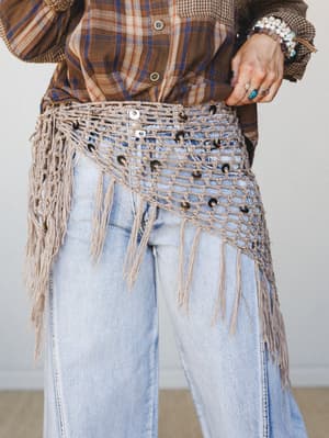 A close-up of a macramé fringe belt layered over light wash wide-leg jeans and a brown plaid shirt, styled with stacked beaded bracelets and rings — earthy, expressive, and very much the kind of thing you'd wear to make a regular outfit feel like a w