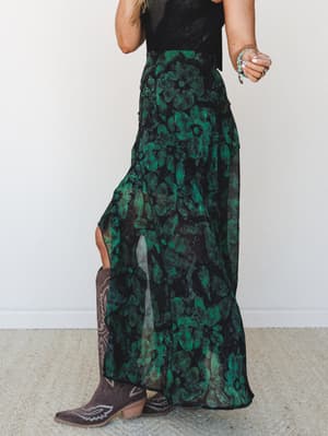 A moody, dramatic maxi skirt in a deep emerald floral print styled with a black top and brown cowboy boots — the kind of outfit that shows up to the concert and stays for the after-party.