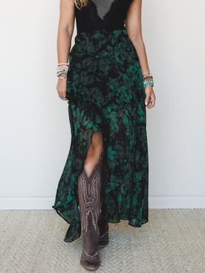 A moody green and black floral maxi skirt with a front slit and sheer chiffon layers, styled with cowboy boots and stacked bracelets for that effortless boho-Western crossover.