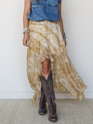A flowy high-low floral skirt in golden yellow pairs with a denim vest and dark cowboy boots — the kind of outfit that looks equally at home at a country concert or a farmers market.