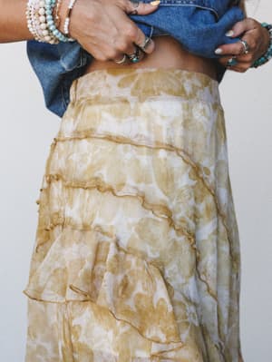 A close-up detail shot of a tiered ruffle skirt in a soft golden floral print, styled with a knotted denim top and layered boho jewelry — the kind of outfit that feels equally at home at a farmers market or a backyard wedding.