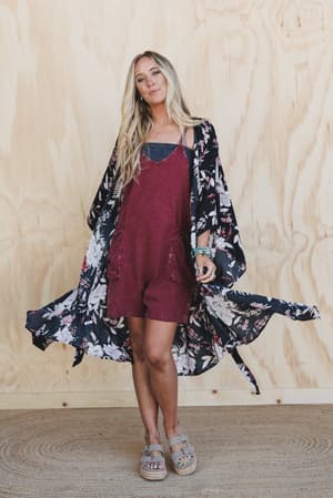 A woman with long blonde hair wears the Free Flowin' Butterfly Sleeve Kimono - Black over a striped top and maroon overalls, paired with sandals. She stands on a woven rug before a light wood background, looking relaxed and stylish.