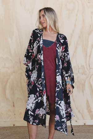 A woman wearing the Free Flowin' Butterfly Sleeve Kimono - Black stands in front of a light wooden wall, looking to the side with one hand in her pocket.