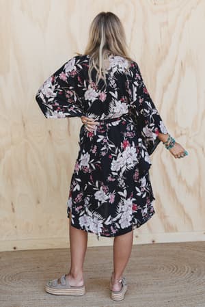 A woman stands with her back to the camera wearing the Free Flowin' Butterfly Sleeve Kimono - Black, paired with platform sandals and turquoise bracelets, set against a light wooden background.