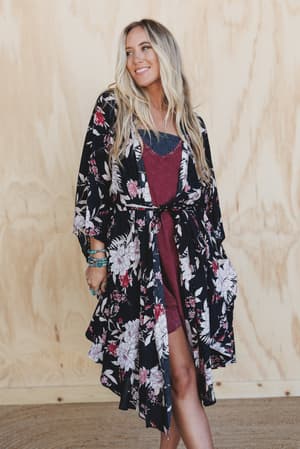 A woman with long blonde hair smiles in a maroon dress and the Free Flowin' Butterfly Sleeve Kimono - Black, featuring a floral pattern. She stands against a light wooden background, accessorized with layered bracelets.