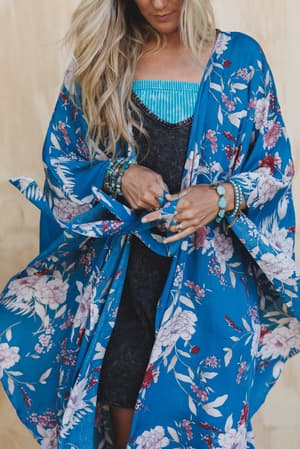 A woman with long blonde hair adjusts the tie on the Free Flowin' Butterfly Sleeve Kimono - Teal over a black dress, wearing turquoise jewelry and a blue bandeau. Her face is partially out of frame.
