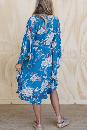 A woman with long blonde hair stands with her back to the camera, wearing the Free Flowin' Butterfly Sleeve Kimono in teal and beige platform sandals. The background has light wood paneling and a textured rug.