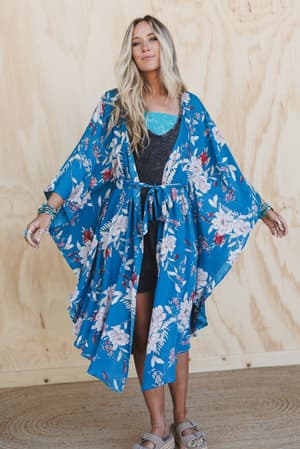 A woman with long blonde hair poses against a light wooden background, wearing the Free Flowin' Butterfly Sleeve Kimono in teal over a black dress and turquoise top, paired with bracelets and sandals, arms slightly outstretched.