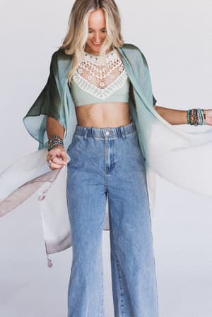 A woman with long blonde hair wears a flowing green and white kimono, the Crochet High Neck Bralette in Sage as a layering piece, high-waisted blue jeans, and colorful beaded bracelets, standing against a plain white background.