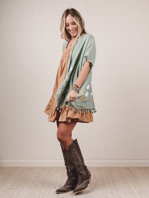 Woman wearing a green dress with ruffled hem, brown skirt, and cowboy boots against a plain background