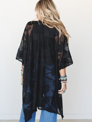 Shot from behind, a model layers a sheer black lace kimono over wide-leg blue jeans, her wrists stacked with boho bracelets — the whole thing feels like a summer evening that turned into something unexpectedly cool.
