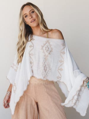 A flowy white embroidered poncho top worn off-shoulder over wide-leg blush pants, styled with stacked beaded bracelets and silver rings — effortless and sun-warmed, like she just stepped off a ferry.